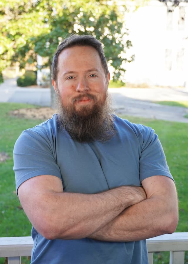 Berea College Information Systems Specialist, Oliver Vaught, leaning against a white fence, with green grass and a tree in the background.