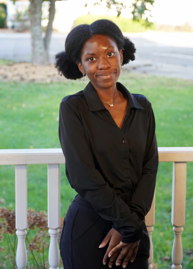 Berea College Admissions Counselor, Yasmina Decamp, standing in front of a white fence, with green grass and a tree in the background.