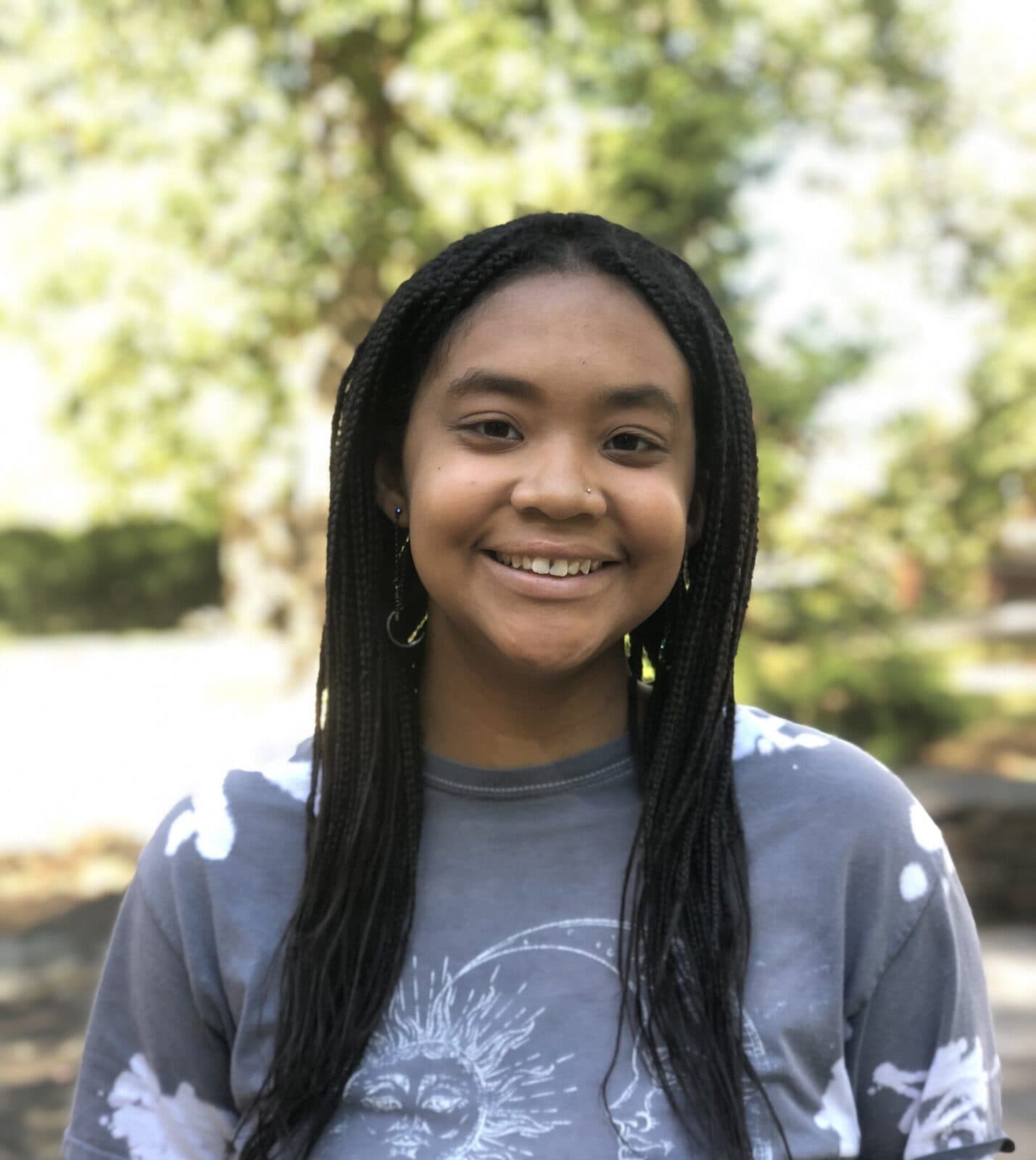 Color image of a Black female in an outdoor setting. The woman has long black braided hair, smiling, and wearing a gray t-shirt with white designs.